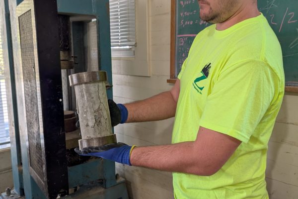 Technician performing compressive strength testing on a concrete cylinder in a laboratory compression machine