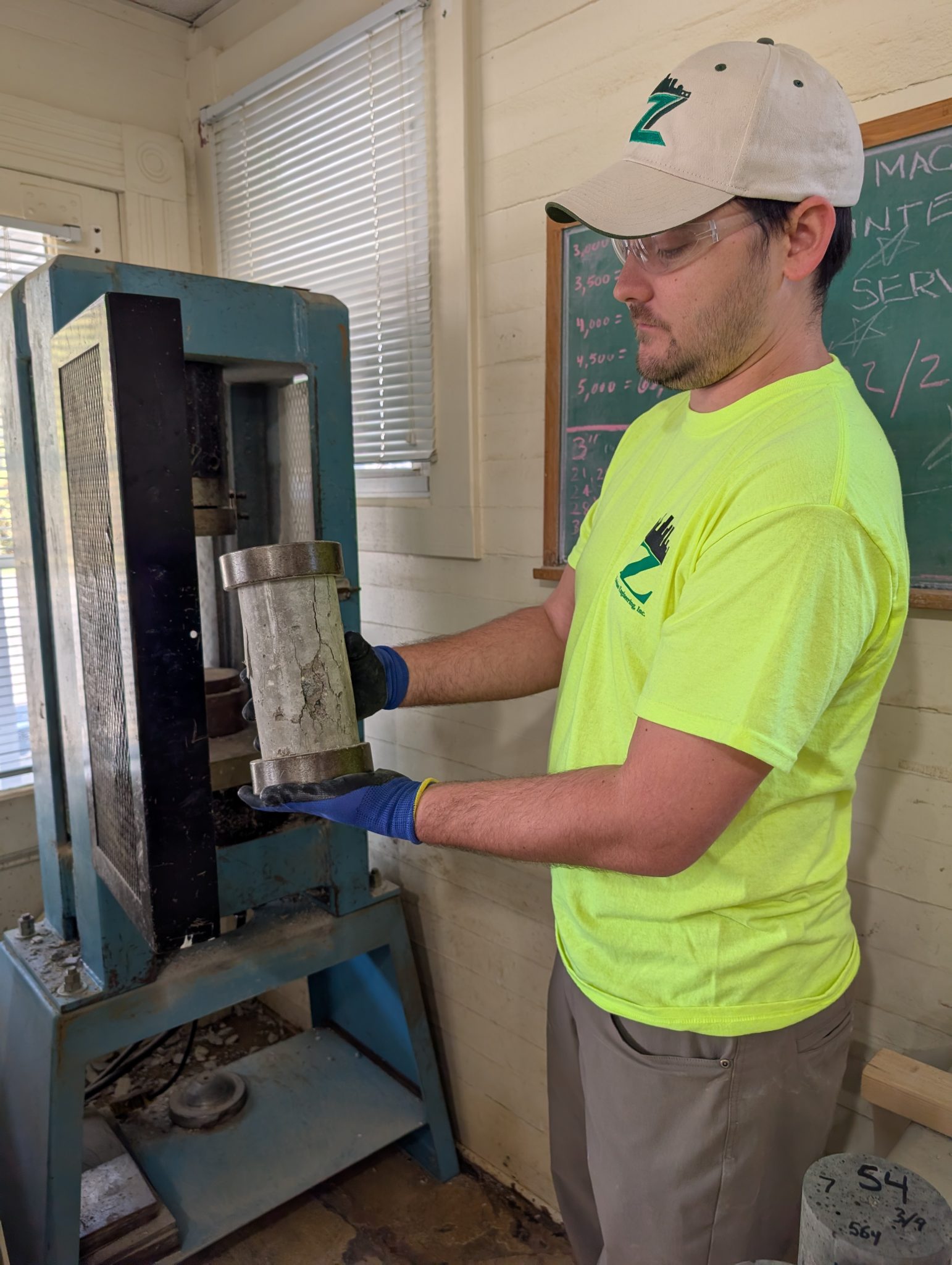 Technician performing compressive strength testing on a concrete cylinder in a laboratory compression machine